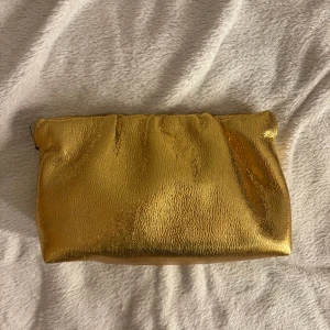 Guldig clutch - Säljer en snygg clutch i metallic guld med mjuk, glansig yta och rynkad öppning. Väskan har svart innerfoder och en liten innerficka. Perfekt för att lyfta din outfit med lite extra bling. Är köpt på mango. Slutsåld på hemsidan!