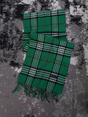 Grön rutig halsduk från Burberrys - Snygg grön halsduk från Burberrys i klassiskt rutigt mönster med inslag av svart, vitt och rött. Tillverkad i 100% lammull och har fransar i ändarna. Perfekt accessoar för att lyfta din outfit med en färgklick och tidlös design.