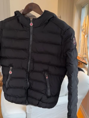 Svart dunjacka med huva från Colmar - Snygg svart dunjacka från Colmar med huva och två dragkedjeförsedda fickor framtill. Jackan har quiltad design och colmar -logga på ärmen. Perfekt för kalla vinterdagar och riktigt lätt att bära.