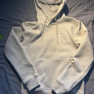 Grå hoodie från Adidas Originals XS - Snygg grå hoodie från Adidas Originals med klassisk logga på bröstet. Tröjan har huva med snörning, magficka och ribbade muddar. Perfekt för chill dagar och streetstyle. Mjuk insida i bomullsmix för extra komfort.