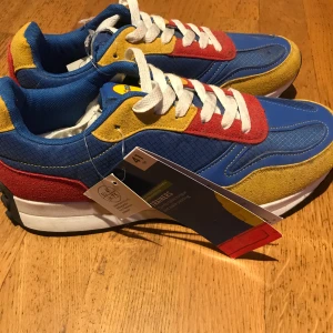 Färgglada Lidl sneakers strl 41 - Säljer ett par unika Lidl sneakers i blått, gult och rött med vit snörning. Skorna har en retro vibe med platt sula, rund tå och coola färgblock. Materialet är en mix av syntet och mocka, och Lidl-loggan syns på plösen. Perfekt för dig som gillar statement pieces!