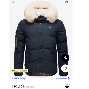 Stone harbor jacka  - Mörkblå pufferjacka från Stone Harbour med stor huva och ljus fuskpäls. Jackan har flera coola detaljer som logopatchar, dragkedjor på ärmen och tryck framtill. Fodrad med mjukt material och har praktiska fickor. Perfekt för kalla dagar och snygg streetstil.