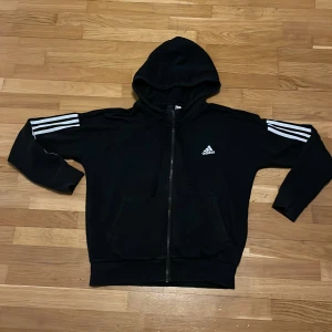 Svart Adidas hoodie med dragkedja - Svart hoodie från Adidas med klassiska vita ränder på ärmarna och logga på bröstet. Tröjan har huva, snörning och hel dragkedja framtill samt två fickor. Perfekt för chill eller träning.