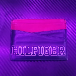 Svart och röd plånbok från Hilfiger - Slimmad plånbok från Hilfiger i skinn med två fack på framsidan och ett större fack i mitten. Färgen är en snygg kombo av röd och svart, med Hilfiger-loggan tryckt i vitt på framsidan. Perfekt för kort och sedlar, och lätt att ha med sig i fickan.