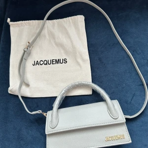 Ljusgrå Jacquemus handväska i skinn - Never used. Supersnygg ljusgrå handväska från Jacquemus med guldfärgad logga framtill. Väskan har en stilren, rektangulär form, justerbar axelrem och ett praktiskt handtag. Materialet är präglat skinn som ger en lyxig känsla. Perfekt för dig som gillar minimalistisk design och vill sticka ut.