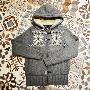 Stickad Vintage hoodie med foder🐻 - Mysig grå stickad hoodie från Alprausch med vitt foder i huvan och snyggt snöflingemönster i vitt och mörkgrått. Tröjan har knappar framtill, snörning vid huvan och ribbade muddar. Perfekt för kalla dagar och chill stunder.