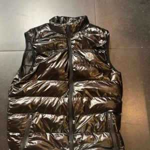 Svart dunväst från Moncler - Snygg svart dunväst från Moncler med glansig finish och klassisk logga på bröstet. Västen har hög krage, två dragkedjefickor och stängs med dragkedja framtill. Perfekt för lager-på-lager och ger en sportig vibe. Pris kan diskuteras vid snabb affär