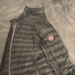 Svart dunjacka från Colmar - Säljer en svart dunjacka från Colmar med klassisk quiltad design och rund Colmar-logga på ärmen. Jackan har dragkedja framtill och en stilren, enkel look som passar perfekt till höst och vinter. Materialet är lätt och smidigt, vilket gör den enkel att bära.