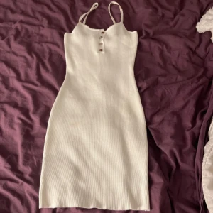 Vit ribbad klänning med knappar - Supersöt vit ribbad klänning med smala axelband och silvriga knappar framtill. Klänningen är figurnära och har en enkel. Materialet är stretchigt och bekvämt. Lite foundation fläckar som försvinner med medel.