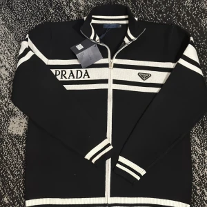 Svart kofta med vita ränder från Prada - Snygg svart kofta från Prada med vita ränder och logotyp på bröstet. Tröjan har hel dragkedja, ribbade muddar och krage. Stora PRADA-bokstäver på framsidan ger en lyxig vibe. Perfekt för dig som gillar stilrena designer.