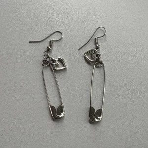 Silvriga örhängen med säkerhetsnål och hjärta - Unika silvriga örhängen formade som säkerhetsnålar med en liten hjärtberlock upptill. Perfekt accessoar för dig som gillar edgy och lekfull stil. Lätta att bära och ger en cool touch till din look.