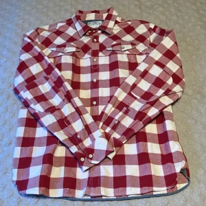 Rödvit rutig Levis skjorta - Snygg röd och vit rutig skjorta från Levi's i storlek L. Skjortan har klassisk westernstil med kontrasttyg i krage och manschetter, bröstfickor med knapp och vita knappar framtill. Tillverkad i mjuk bomull, perfekt för dig som gillar retrovibbar och coola detaljer.