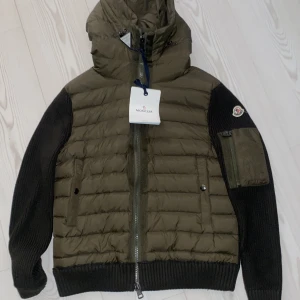 Olivgrön Moncler dunjacka med stickade ärmar - Snygg olivgrön dunjacka från Moncler med stickade svarta ärmar och huva. Jackan har dragkedja framtill, fickor med knappar och en extra ficka på ärmen med Moncler-logga. Perfekt för dig som vill ha en stilren och varm jacka med exklusiv känsla.