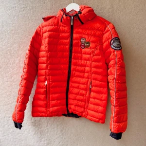 Röd pufferjacka från Napapijri L - Klarröd / korallröd dunjacka från Napapijri modell ’Armes down’.  Avtagbar luva, sidofickor, innerficka, dragkedja fram.  Ytterfoder i 100% polyamid; ett extremt slitstarkt och tåligt material. Vattenavvisande, snabbtorkande och dessutom superskönt att bära.  Stoppningen består av 80% anddun och 20% dunfjädrar. Fläck på höger arm. I övrigt fint skick. Storlek L Bröstvidd ~104cm Ärm ~ 50cm Längd ~65cm