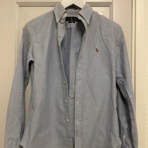Blå skjorta från Ralph Lauren - Snygg ljusblå skjorta från Ralph Lauren med klassisk broderad logga på bröstet. Jag har haft denna skjortan ett tag men har bara använt den på speciella sammanhang så inte så använda. Hör gärna av dig vid frågor