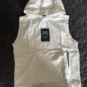 Vit ärmlös hoodie-väst från Zara - Cool vit hoodie-väst från Zara med tryckt patch framtill. Västen har stor huva, magficka och dragkedja i sidan. Perfekt för en sportig look och enkel att matcha med andra plagg. Tillverkad i mjuk bomull för skön känsla.