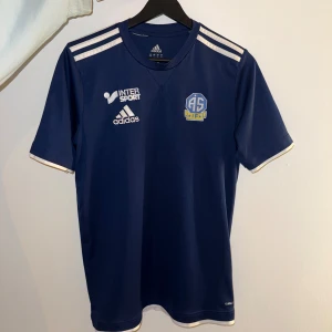 Blå Adidas fotbollströja  - Snygg marinblå fotbollströja från Adidas, vita detaljer och klassiska Adidas-ränder på axlarna. Tillverkad i lätt och ventilerande Climalite-material, perfekt för träning eller match.
