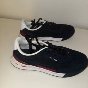 Mörkblå sneakers från Tommy Hilfiger - Snygga mörkblå sneakers från Tommy Hilfiger med vita och vinröda detaljer. Skorna har svart snörning, vit innersida och en platt sula. Materialmix av mocka och syntet ger en sportig och stilren look. Perfekta för dig som gillar klassisk streetstyle.