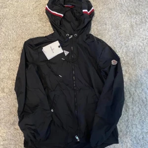 Svart vindjacka från Moncler med huva - Svart vindjacka från Moncler med huva och dragsko, klassisk logga på ärmen och snygga rödvita detaljer på huvkanten. Jackan har dragkedja framtill och praktiska fickor med tryckknapp. Perfekt för dig som vill ha en stilren och sportig look.