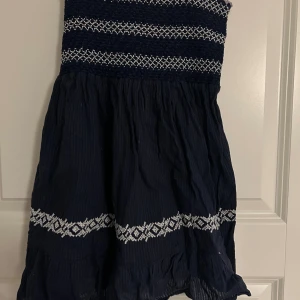 Marinblå långt linne från Vila XS - Supersöt marinblå långt linne från Vila med smockad överdel och tunna axelband. Linnet har vita broderade detaljer och volangkant nertill. Perfekt för sommarens alla tillfällen och ger en fräsch och ungdomlig look.