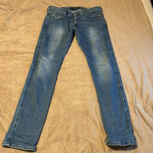 Replay Anbass blå jeans, storlek 32 - Snygga Replay Anbass jeans i klassisk blå tvätt med lätt slitningar och raka ben. Jeansen har fem fickor, knappstängning och coola detaljer på bakfickorna. Perfekt passform och stilren look för dig som gillar street och casual.