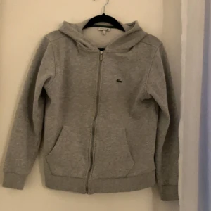 Grå hoodie från Lacoste med dragkedja - Grå zip hoodie från Lacoste. Bra skick. Storlek 12.