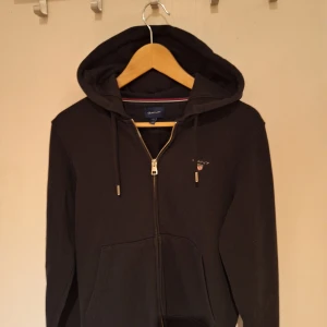 Svart hoodie från GANT med dragkedja - Svart hoodie från GANT med huva, snörning och hel dragkedja framtill. Klassisk logga broderad på bröstet och stora fickor framtill. Perfekt för chill dagar och enkel att matcha med jeans eller joggers.