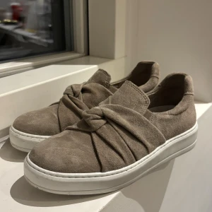 Beige mocka sneakers med rosett - Snygga beige sneakers från VICOBLER i mjuk mocka med en unik rosettdetalj över foten. Skorna har platt vit sula och rund tå, vilket ger en clean och modern look. Perfekta för dig som gillar stilrena och bekväma skor med en twist.