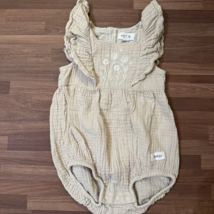 Beige romper - Oanvänd och i nyskick. Supersöt beige romper från Newbie i storlek 56 (1-2mån). Volangdetaljer på axlarna och broderade blommor framtill. Träknappar i ryggen samt tryckknappar i grenen för enkel påklädning.