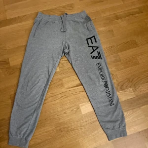 Grå mjukisbyxor EA7 Emporio Armani - Grå mjukisbyxor från EA7 Emporio Armani med svart logga längs benet och på bakfickan. Byxorna har resår och snörning i midjan samt muddar vid bensluten. Tillverkade i mjuk bomullsmix för maximal komfort.