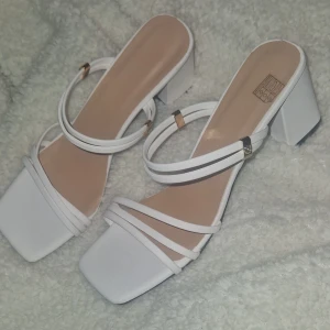 Klackskor - Snygga vita helt nya klackskor. Skorma har bara legat i ett skåp, och måste därför ha blivit en liten repa på ena klacken. Skorna har tunna remmar över foten och runt vristen med små gulddetaljer. Perfekta för nästintill alla outfits och ger en fräsch look. 