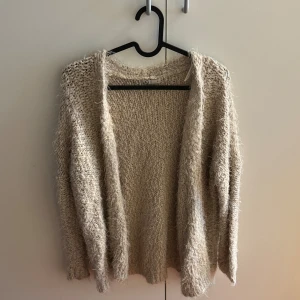 Beige fluffig stickad kofta från Cubus - Mysig och fluffig beige stickad kofta från Cubus i storlek S. Koftan har långärmade ärmar och är öppen framtill utan knappar. Den grova stickningen och de fluffiga trådarna ger en chill och avslappnad vibe, perfekt för lager-på-lager under kyliga dagar.
