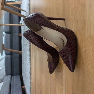 Vinröda pumps med ormskinnsmönster - Snygga vinröda pumps med hög klack. Skorna har ett ormskinnsmönstrat yttre i glansigt material som ger en lyxig känsla. Perfekta för dig som vill sticka ut med en elegant och trendig look.