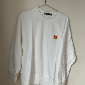 Vit sweatshirt från Acne Studios - Vit sweatshirt från Acne Studios med klassisk rund hals och lång ärm. På bröstet sitter den ikoniska orangea broderade face-patchen. Tröjan har en loose passform och är tillverkad i mjuk bomull som är skön mot huden.