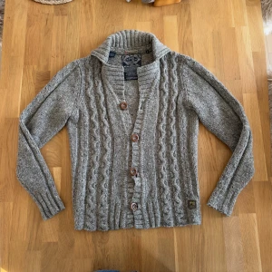 Grå stickad kofta från Scotch & Soda - Säljer en grå stickad kofta från Scotch & Soda med snyggt flätmönster och grov ribbad krage. Koftan har fyra bruna knappar framtill och är långärmad. Perfekt för dig som gillar chill och stilren look under höst och vinter.