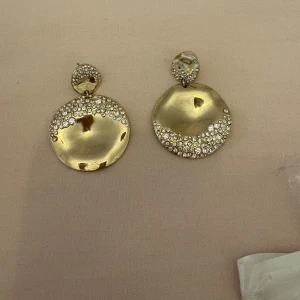 Stora guldiga örhängen med strass - Säljer ett par stora, runda örhängen i guldig metall med glittrande strassdetaljer på både den lilla och stora cirkeln. De har en cool, vågig yta och ger en riktigt statement-look. Perfekta för dig som vill sticka ut och älskar bling!