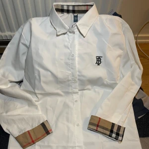 Vit Burberry-inspirerad skjorta - Vit skjorta med klassisk krage och knäppning framtill. Har Burberrys ikoniska rutiga mönster på insidan av kragen och ärmsluten samt broderad logga på bröstet. Skjortan är långärmad och i bomull, perfekt för en clean och stilren look.