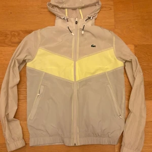 Beige vindjacka från Lacoste med huva - Snygg beige vindjacka från Lacoste med ljusgul detalj över bröstet, dragkedja framtill och justerbar huva. Jackan har elastiska muddar och två fickor med dragkedja. Perfekt för dig som vill ha en stilren och sportig look.