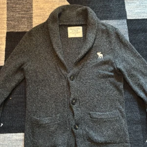 Mörkgrå kofta Abercrombie & Fitch - Mysig mörkgrå kofta från Abercrombie & Fitch med två framfickor och klassisk sjalkrage. Tröjan har långa ärmar, knäppning med tre knappar och en vit broderad älg på bröstet. Perfekt för lager-på-lager och chill stil. Aldrig använd 