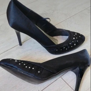 Svarta pumps med klack och pärldetaljer - Snygga svarta klackar från Naomi i storlek 40. Skorna har spetsig tå, hög klack och är dekorerade med små pärlor framtill. Materialet har en satinliknande finish som ger en elegant look. Perfekt för formella event. Klackhöjd: 9,5 cm. Innersula: 27 cm