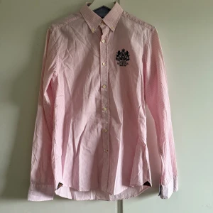 Rosa randig skjorta från Morris - Säljer en klassisk rosa och vit randig skjorta från Morris med broderad logga på bröstet. Skjortan har lång ärm, knappar framtill och button down-krage. Perfekt för dig som gillar preppy stil och snygga detaljer.
