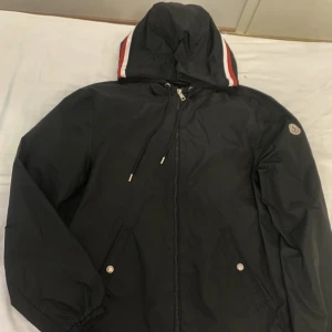 Svart vindjacka från Moncler med huva - Svart vindjacka från Moncler med huva och dragkedja framtill. Jackan har Moncler-logga på ärmen. Nästan helt ny. Äkta såklart köpt på Plick. Sälja pågrund av att jag har bytt stil så har inte behov av den längre 