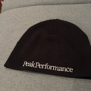 Svart mössa Peak Performance - Svart, stilren mössa från Peak Performance med vit broderad logga framtill. Klassisk rundad form och ribbad stickning som ger en snygg look. Perfekt för kalla dagar och enkel att matcha med din outfit.
