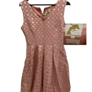 Rosa kortklänning Maria Westerlind - Superfin rosa kortklänning från Maria Westerlind med silvriga blommönster och v-ringning fram. Klänningen har markerad midja och veckad kjol för extra volym. Limited edition och exklusivt material – perfekt för dig som vill sticka ut.