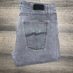 Nudie jeans  - Snygga grå jeans från Nudie Jeans med klassisk femficksdesign och svarta detaljer på bakfickorna. Jeansen har en smal passform och raka ben, tillverkade i mjukt denimtyg. Midja 43cm, längd 109cm. Storlekslapp saknas men jag bedömer storleken till W34 L34 men utgå från måtten för att vara säker. OBS liten fläck på höger sida utav höger ficka JAG SKICKAR INGA FLER BILDER NÄR JAG BÄR JEANSEN PÅ :) s2 