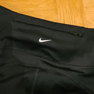 Nike Dri-Fit svarta leggings XS - Svarta Nike Dri-Fit leggings i storlek XS med hög midja och tight passform. Byxorna har diskret Nike-logga framtill och en liten dragkedjeficka bak. Tillverkade i ett stretchigt syntetmaterial som andas, perfekta för träning.
