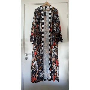 Kimono / klänning  - Helt NY flerfärgad kimono i strl L med fjärilsmönster i svart, vitt, orange och brunt. Kimonon har en svartvit randig kant längs öppningen och ärmsluten. Lika snygg över klänning / jeans eller som klänning med bälte i midjan.   Hämtas i Stockholm eller skickas mot porto. 