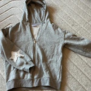 Grå hoodie med stjärndetalj - Grå hoodie med dragkedja och huva. Tröjan har en stor vit stjärna på ryggen och en mindre stjärna på ena ärmen. Mjuk och skön med ribbade muddar och klassisk passform. Perfekt för chill dagar eller när du vill sticka ut lite extra.
