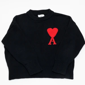 Svart stickad tröja från Ami Paris - Svart stickad tröja från Ami Paris med rund halsringning och ett rött hjärta med bokstaven A broderat på bröstet. Tröjan har långa ärmar och breda ribbade muddar vid ärmslut och nederkant. Perfekt för dig som gillar stilrena detaljer med en twist. Det finns ett litet streck precis ovanför loggan som syns vid noga inspektion.