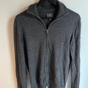 Mörkgrå Merinoull kofta med dragkedja från H&M - Mörkgrå kofta från H&M med hel dragkedja och hög krage. Tröjan är långärmad och har en stilren, enkel design som passar perfekt till lager-på-lager. Tillverkad i mjuk bomullsblandning för extra komfort.